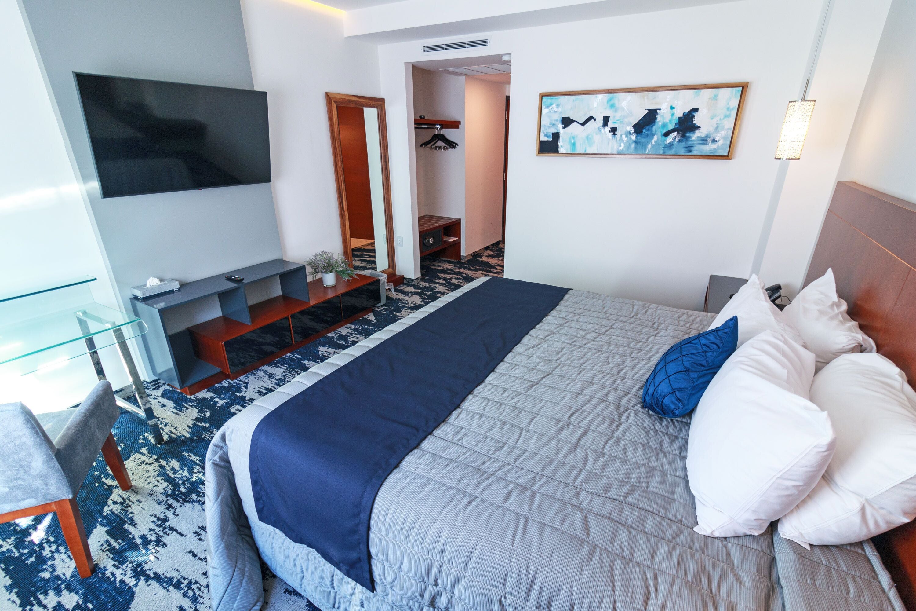 Quarto de hotel com uma cama grande, uma televisão de tela plana na parede, uma área de estar com mesa de vidro e um corredor que leva a um banheiro.