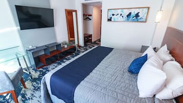 Habitación de hotel con una cama grande, un televisor de pantalla plana en la pared, una zona de estar con una mesa de vidrio y un pasillo que conduce a un baño.