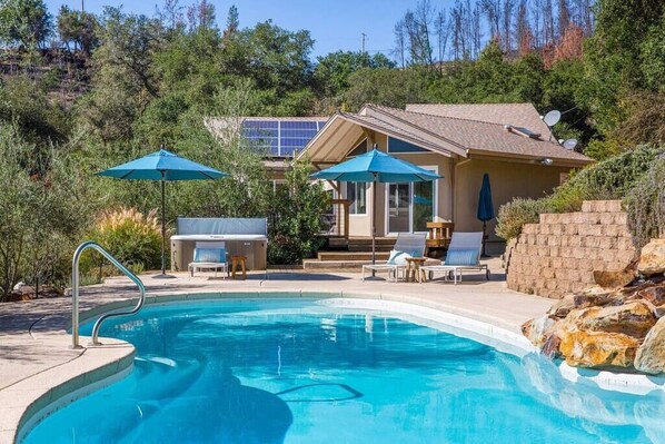 Piscina con sillas de descanso y sombrillas, rodeada por una casa con paneles solares y vistas a árboles.