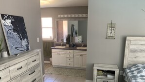 A modern bathroom with a vanity, sink, and mirror. There is a small bedside table with a lamp and a bed with a patterned blanket. - Casa Tranquilo    Heated Pool and Sauna (Gulf Breeze)