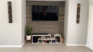 A living room with a flat-screen TV mounted on a wooden wall, a white entertainment unit below, and a potted plant to the left. - Casa Tranquilo    Heated Pool and Sauna (Gulf Breeze)
