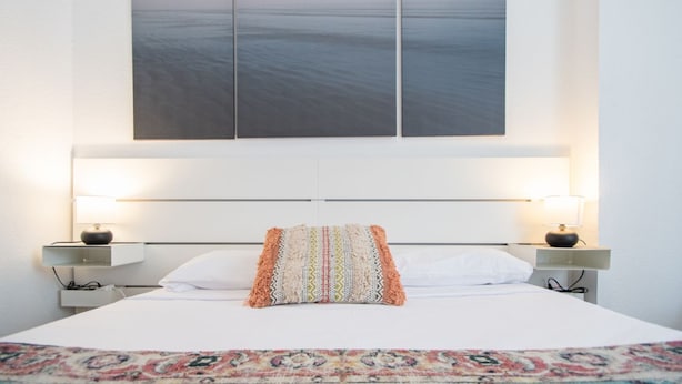 Une chambre d’hôtel avec un grand lit, deux tables de chevet avec des lampes, un plaid à motifs et une œuvre d’art accrochée au mur représentant un paysage marin avec des oiseaux.