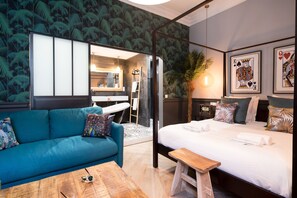 Una habitación de hotel moderna con un sofá color turquesa, una mesa de centro de madera, una cama con ropa blanca y un espejo grande.