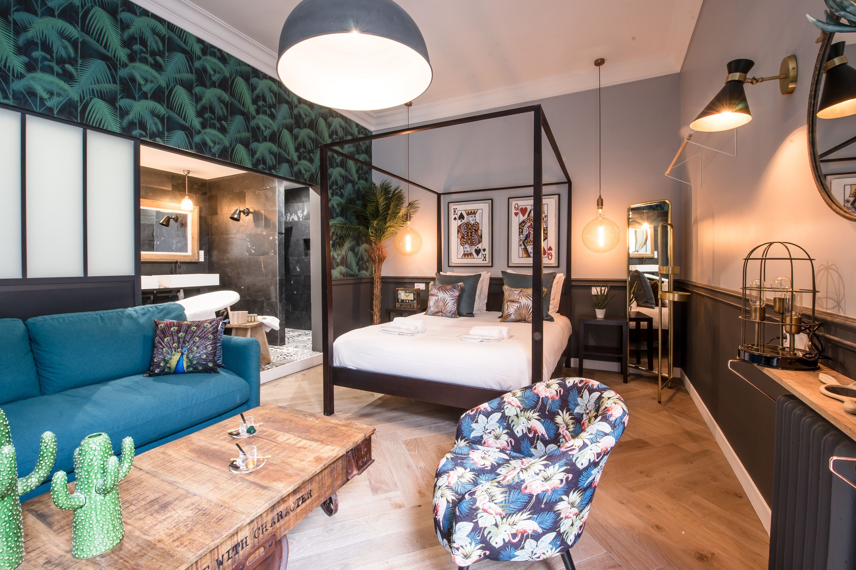 A modern bedroom with a four-poster bed, a blue sofa, a floral armchair, and a wooden coffee table.