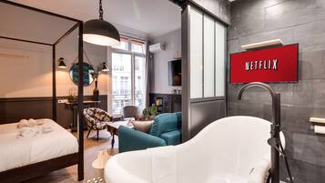 A modern hotel room with a large bed, a freestanding bathtub, a flat-screen TV, and a view of the city.