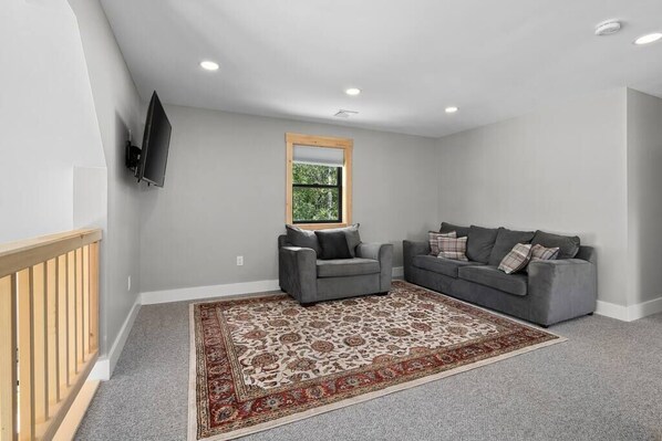 A living room with a grey sofa, a grey armchair, a large patterned rug, a wooden-framed window, and a mounted TV. - Encore Escape | Walk To The Lifts @ Mt. Snow! (Dover)