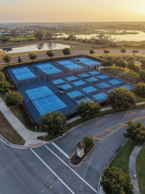 An aerial view of a sports complex with multiple tennis courts, surrounded by greenery and a parking area.