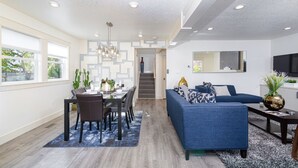 A modern living room with a blue sofa, dining area, and staircase leading to an upper level. - Full House ➡️ Hut Tub (Aloha)