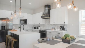 Une cuisine moderne avec des meubles blancs, un îlot central doté de tabourets, une cuisinière et une grande fenêtre.