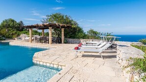 Ein Poolbereich mit Liegestühlen, einer Pergola und Blick auf das Meer.