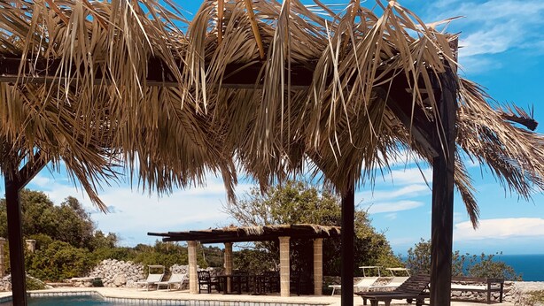 Ein Poolbereich mit einer strohgedeckten Struktur, Liegestühlen und Blick auf das Meer.