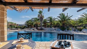 Ein Essbereich am Pool mit einem Tisch für zwei Personen, mit Blick auf einen von Palmen gesäumten Pool und eine Steinmauer.