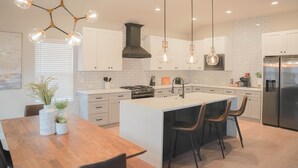 A modern kitchen with white cabinets, a central island with bar stools, a stainless steel refrigerator, and pendant lighting.