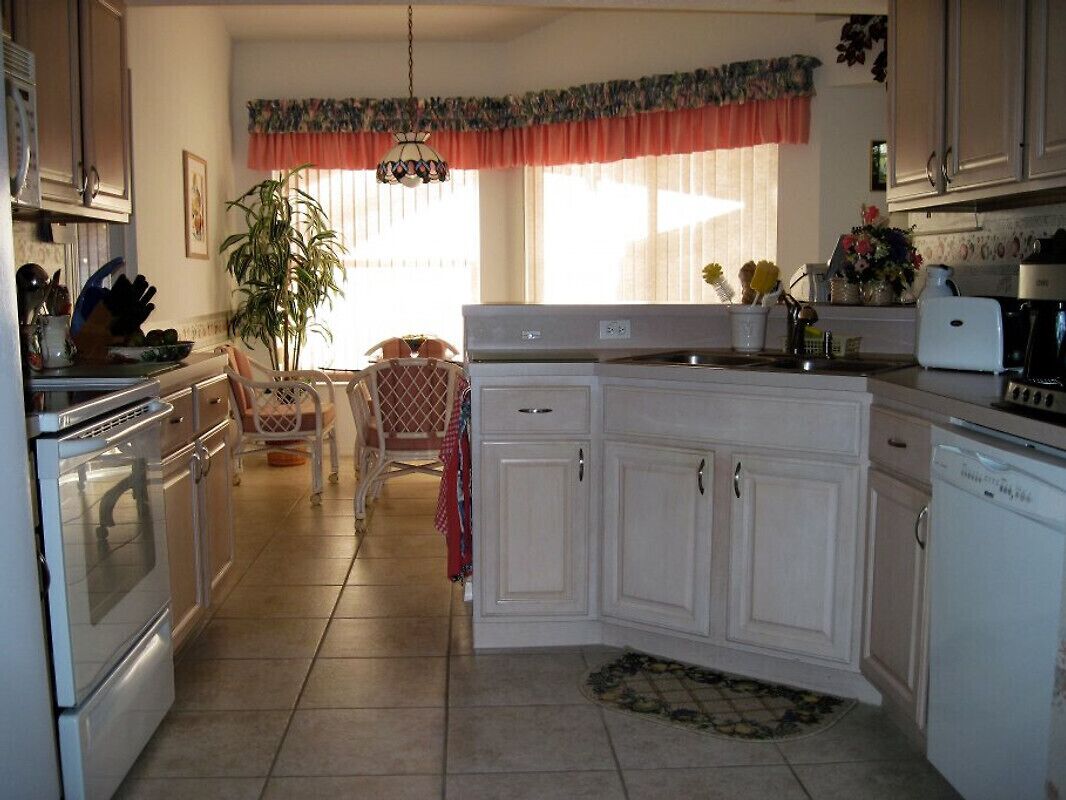 Una cocina con armarios blancos, una estufa, un lavaplatos y un fregadero. Hay una mesa de comedor con sillas y una planta en una esquina.
