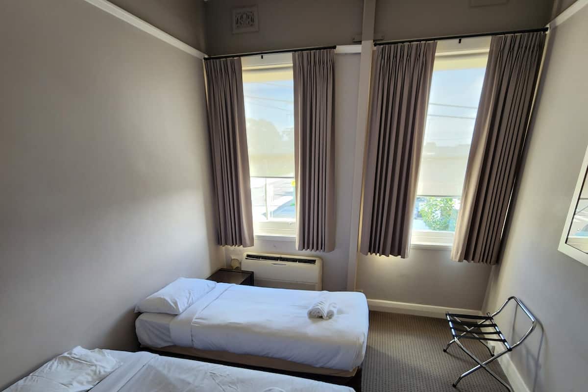 Habitación de hotel con dos camas individuales, un radiador, una ventana con cortinas y una silla fijada a la pared, además de suelo alfombrado.