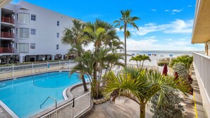 Zona de piscina con vistas despejadas a la playa, rodeada de palmeras y un edificio blanco con balcones.