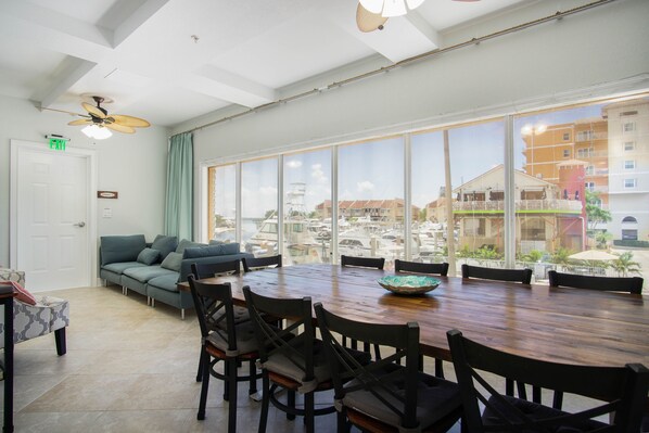 Une pièce spacieuse avec une grande table à manger, des sièges confortables, un ventilateur de plafond et une vue sur une marina, grâce à de grandes fenêtres.