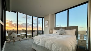 Un dormitorio espacioso con una cama grande, una silla y vistas de la ciudad a través de amplias ventanas.