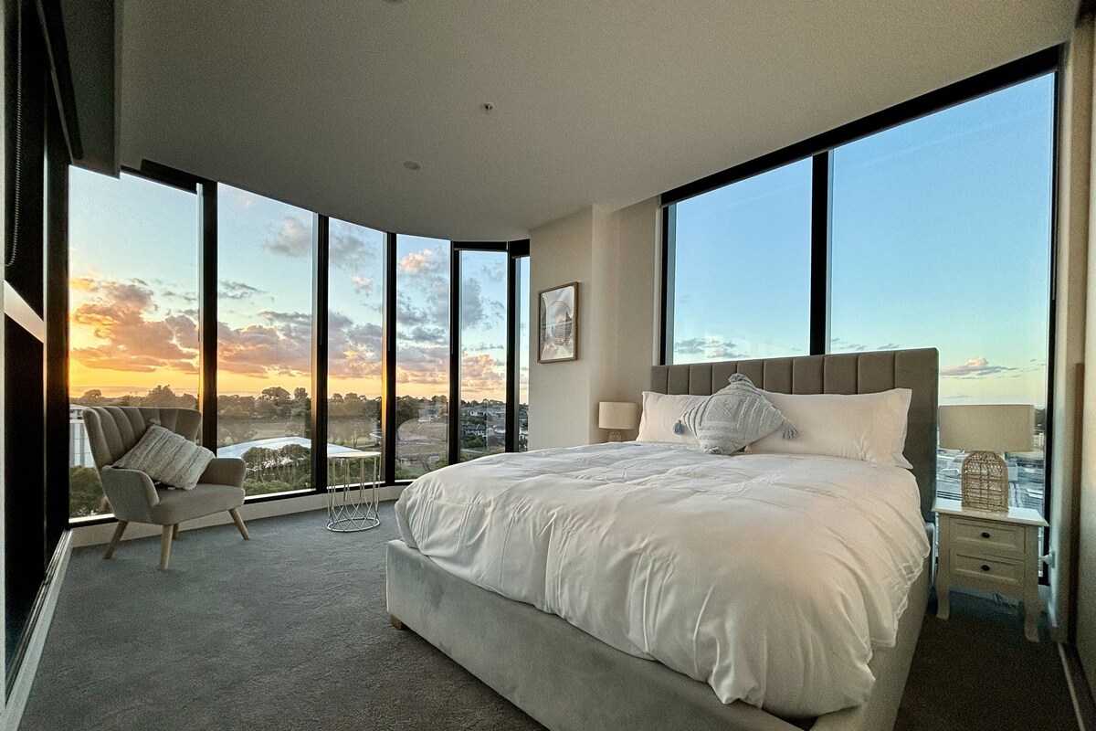 A spacious bedroom with a large bed, a chair, and a view of the cityscape through large windows.