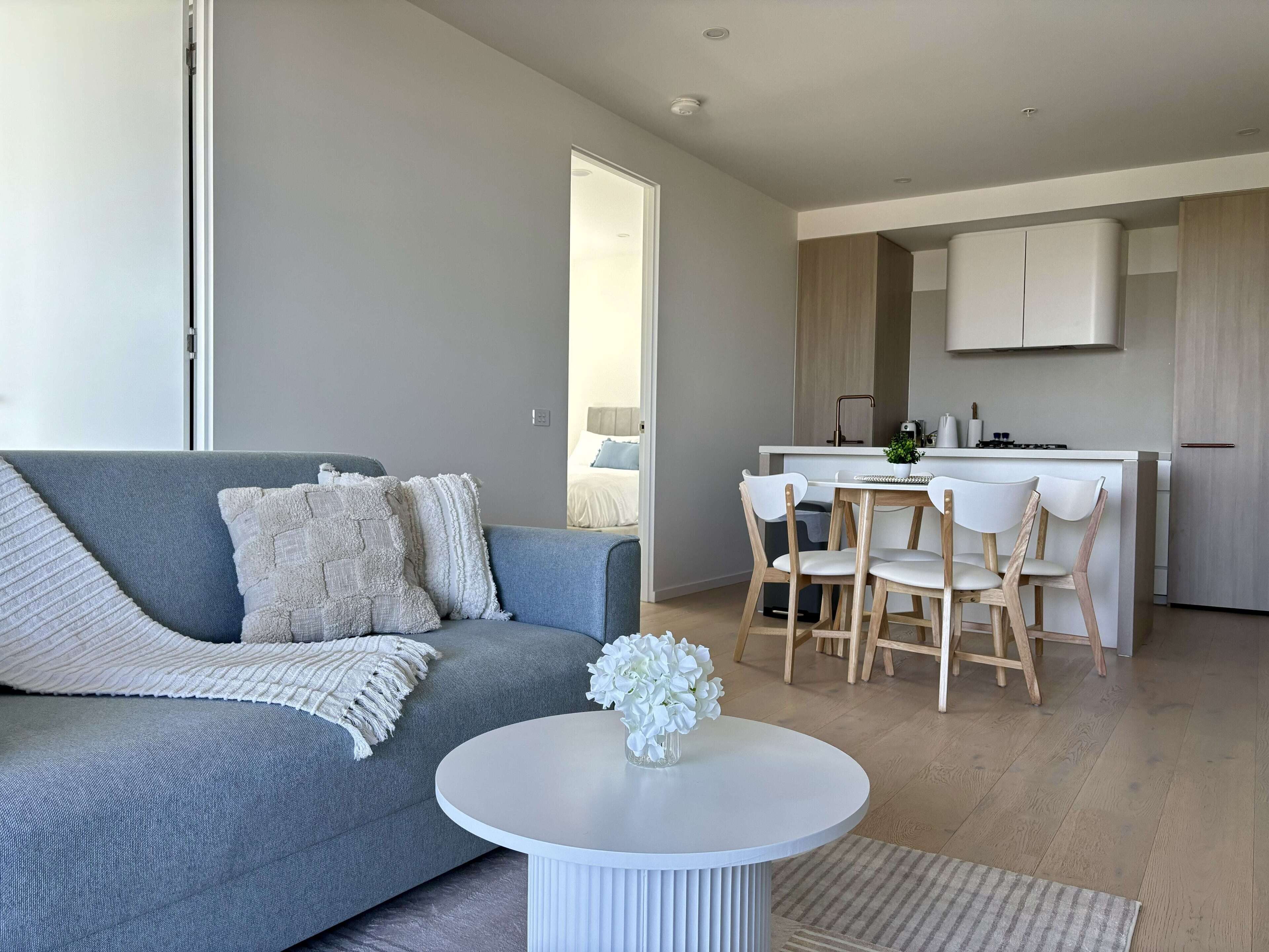 A modern living room with a grey sofa, a round white coffee table, and a dining area with wooden chairs and a small kitchen in the background.