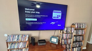 Un televisor mostrando un mensaje de bienvenida, un mueble de madera para entretenimiento con un reproductor de DVD y dos estanterías llenas de DVD.