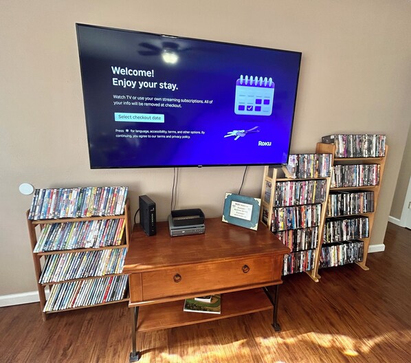 Un televisor mostrando un mensaje de bienvenida, un mueble de madera para entretenimiento con un reproductor de DVD y dos estanterías llenas de DVD.