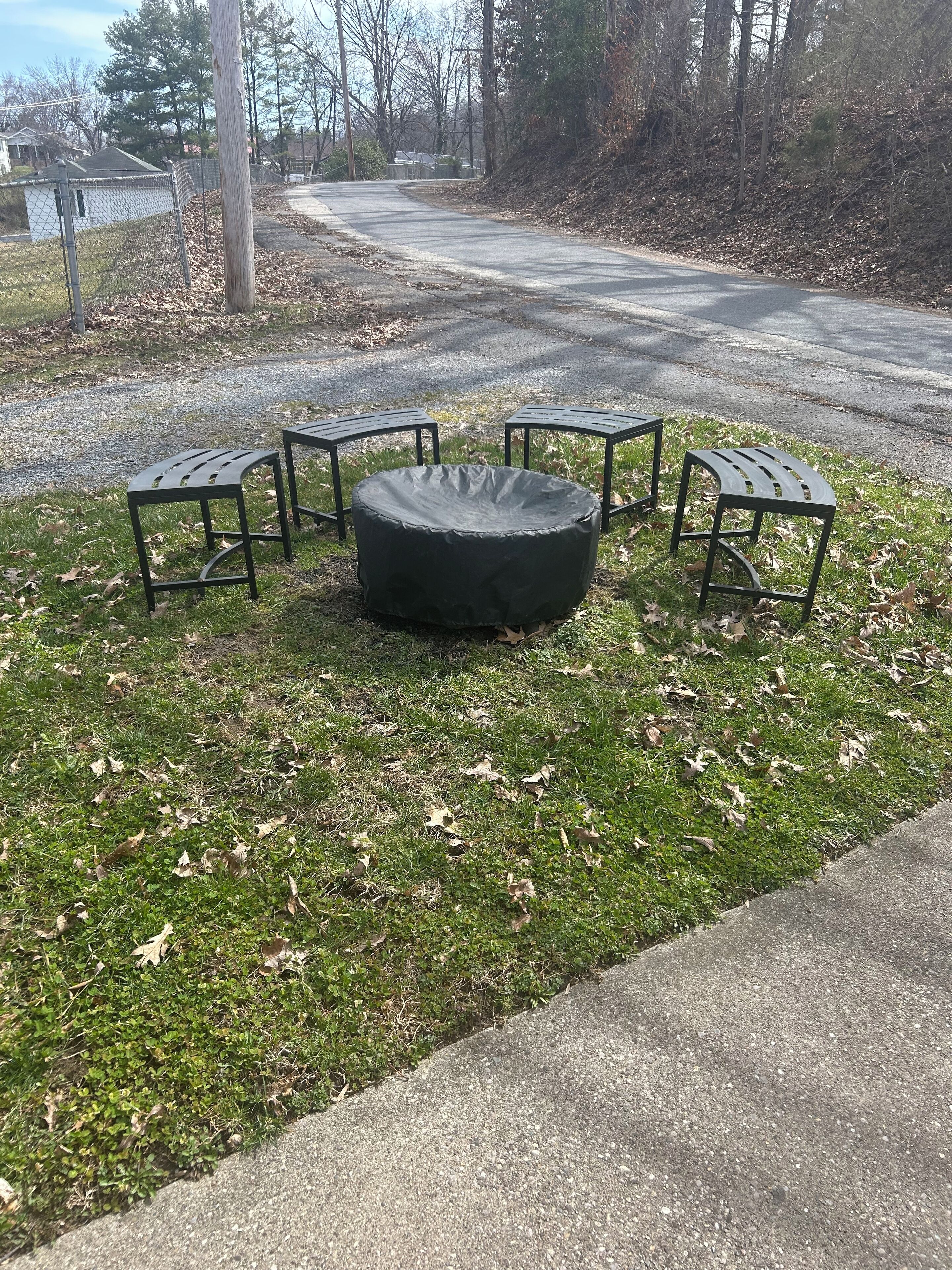 A circular outdoor seating area with four metal tables and benches, surrounded by grass and scattered leaves.
