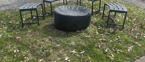 A circular outdoor seating area with four metal tables and benches, surrounded by grass and scattered leaves.