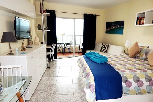 A hotel room with a bed, a desk, a chair, a TV, and a balcony with a table and chairs.