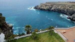 A coastal view with a rocky cliff, a clear blue sea, and a grassy area with a pathway leading to a seating area.