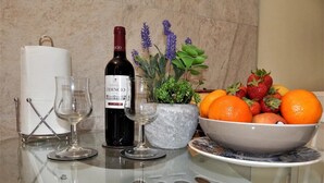 A glass table with a wine bottle, two wine glasses, a bowl of fruit, and a potted plant.