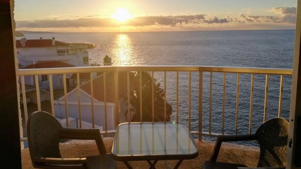 A balcony with two chairs and a small table, overlooking a sunset over the sea with buildings in the background.