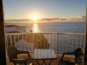 Un balcón con dos sillas y una mesita, con vista a una puesta de sol sobre el mar y edificios al fondo.