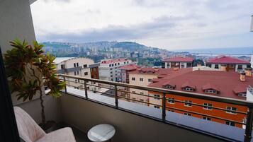 A balcony with a chair and a small table, overlooking a cityscape with buildings and a distant view of the sea.