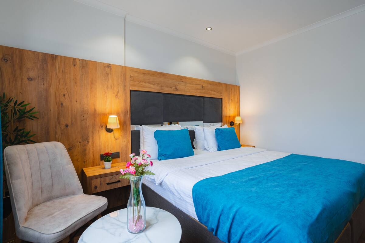 Ein Hotelzimmer mit Holz-Kopfteil, einem Bett mit blauer Bettwäsche, einem Nachttisch mit Lampe, einem Sessel und einer Vase mit Blumen.