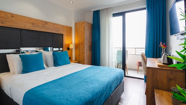Habitació d'hotel amb un llit gran, un capçal de fusta, escriptori amb cadira, televisió i un balcó amb cortines blaves.