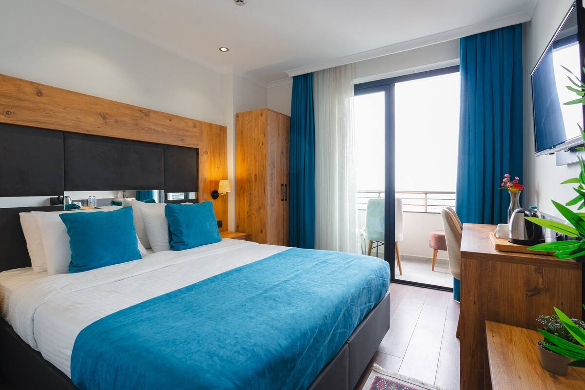 Ein Hotelzimmer mit einem großen Bett, einem Holzkopfende, einem Schreibtisch mit Stuhl, einem Fernseher und einem Balkon mit blauen Vorhängen.