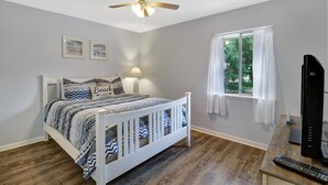 Ein Schlafzimmer mit einem Bett, Deckenventilator, einem Fenster mit Vorhängen, einem Fernseher und Holzboden.