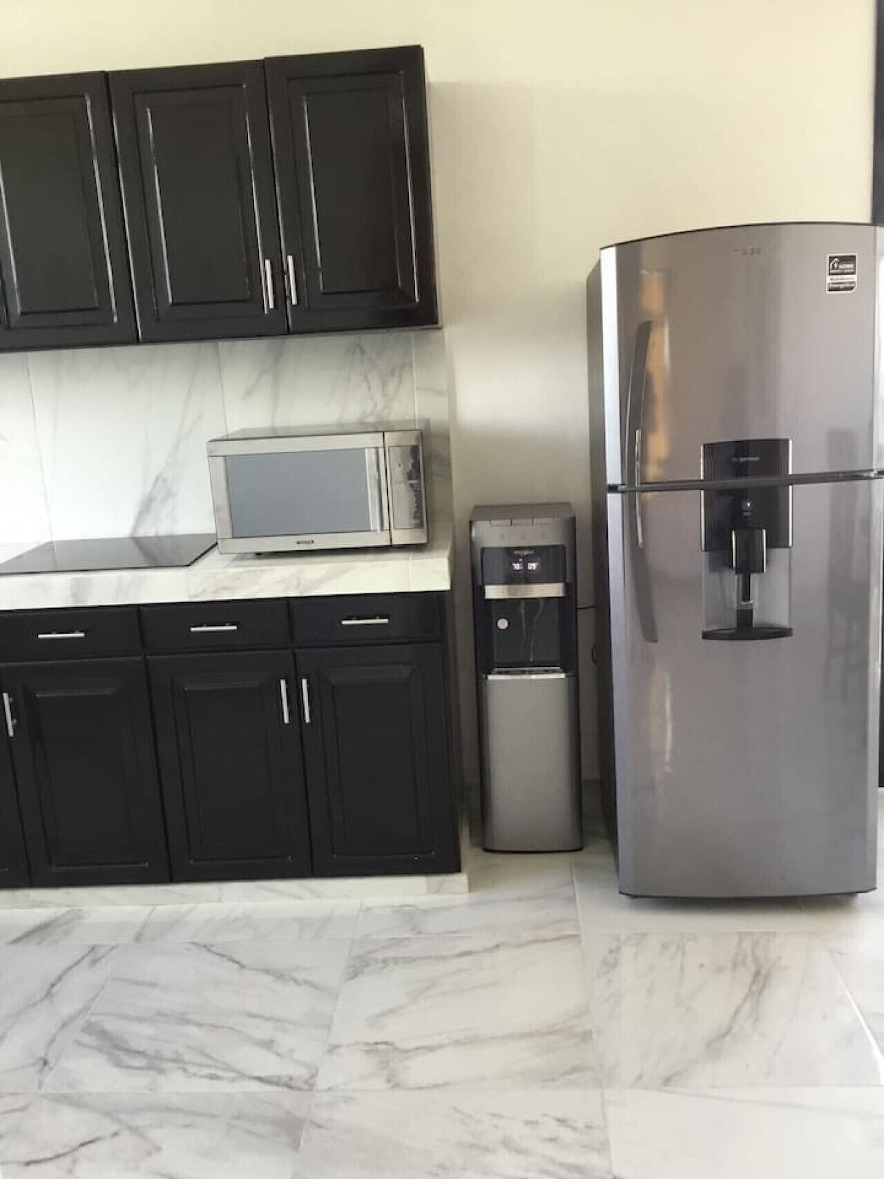 A kitchen with black cabinets, a stainless steel refrigerator, a microwave, and a water dispenser.
