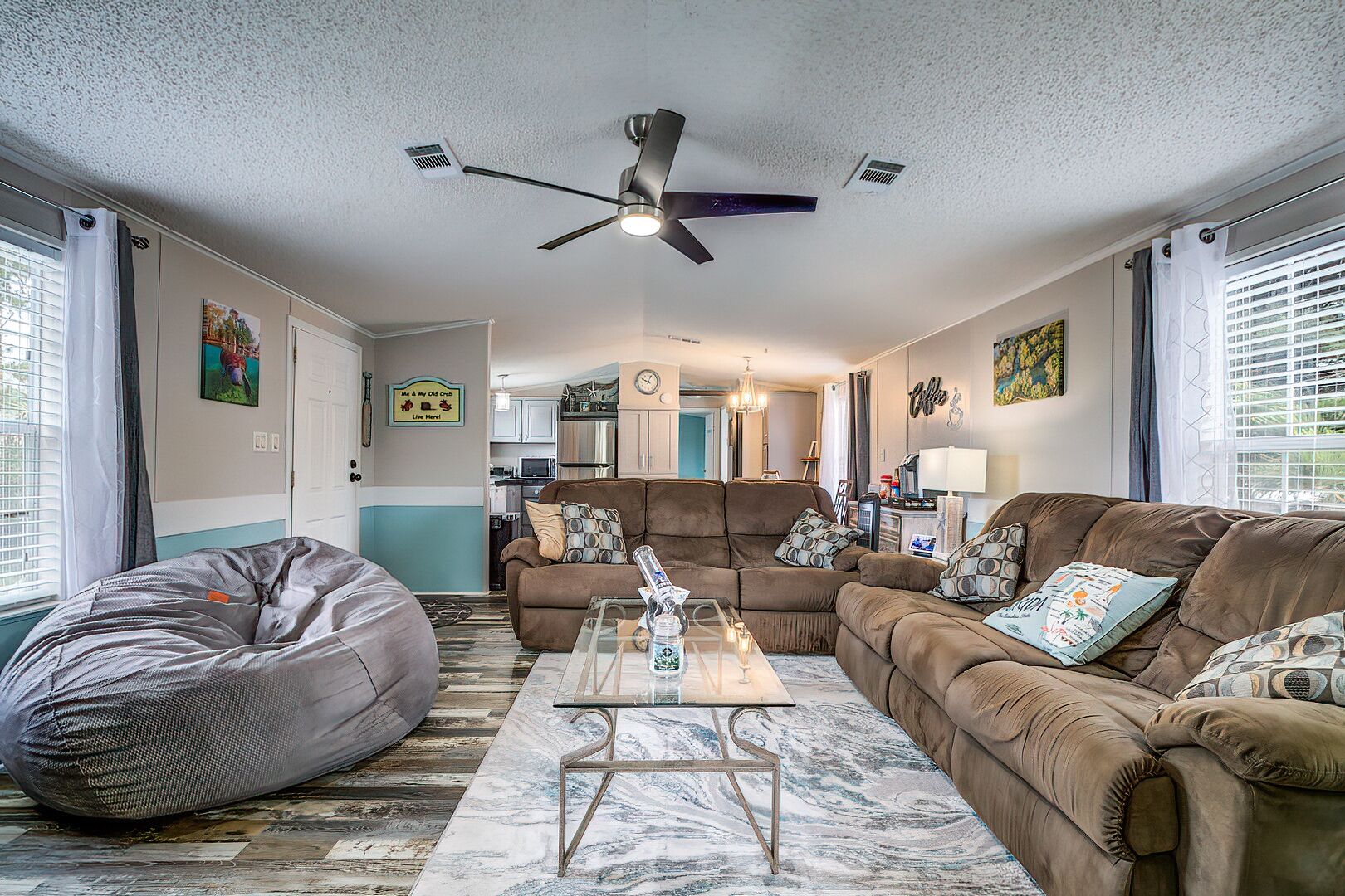 Ein Wohnzimmer mit braunem Sofa, Sitzsack und Glastisch. Ein Deckenventilator ist vorhanden und im Hintergrund befindet sich eine Küche.