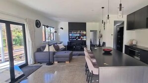 Una cocina moderna con una isla central, un comedor con una mesa larga, y una sala de estar con un sofá y televisor.