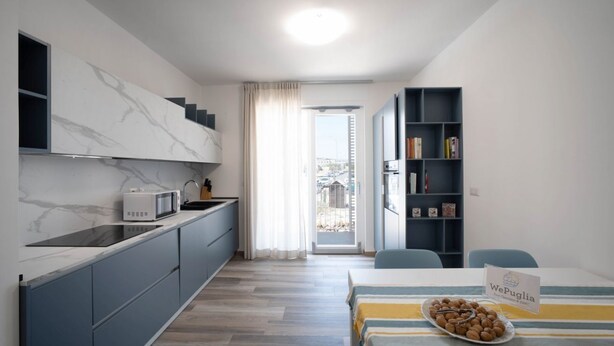 A modern kitchen with a microwave, a dining area with a table set for a meal, and a bookshelf. - WePuglia – Appartamento GiuGio (Polignano a Mare)