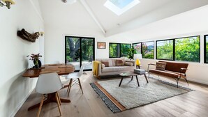 A spacious living room with a high ceiling, large windows, a wooden dining table, and a cozy seating area.