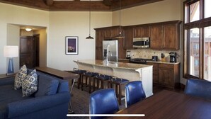 A modern kitchen with wooden cabinets, a stainless steel refrigerator, and a built-in microwave. There is a dining area with a table and chairs, and a living room with a sofa and cushions. - Spring Ski Week in the Canyons- Ski In/Ski Out (Park City)