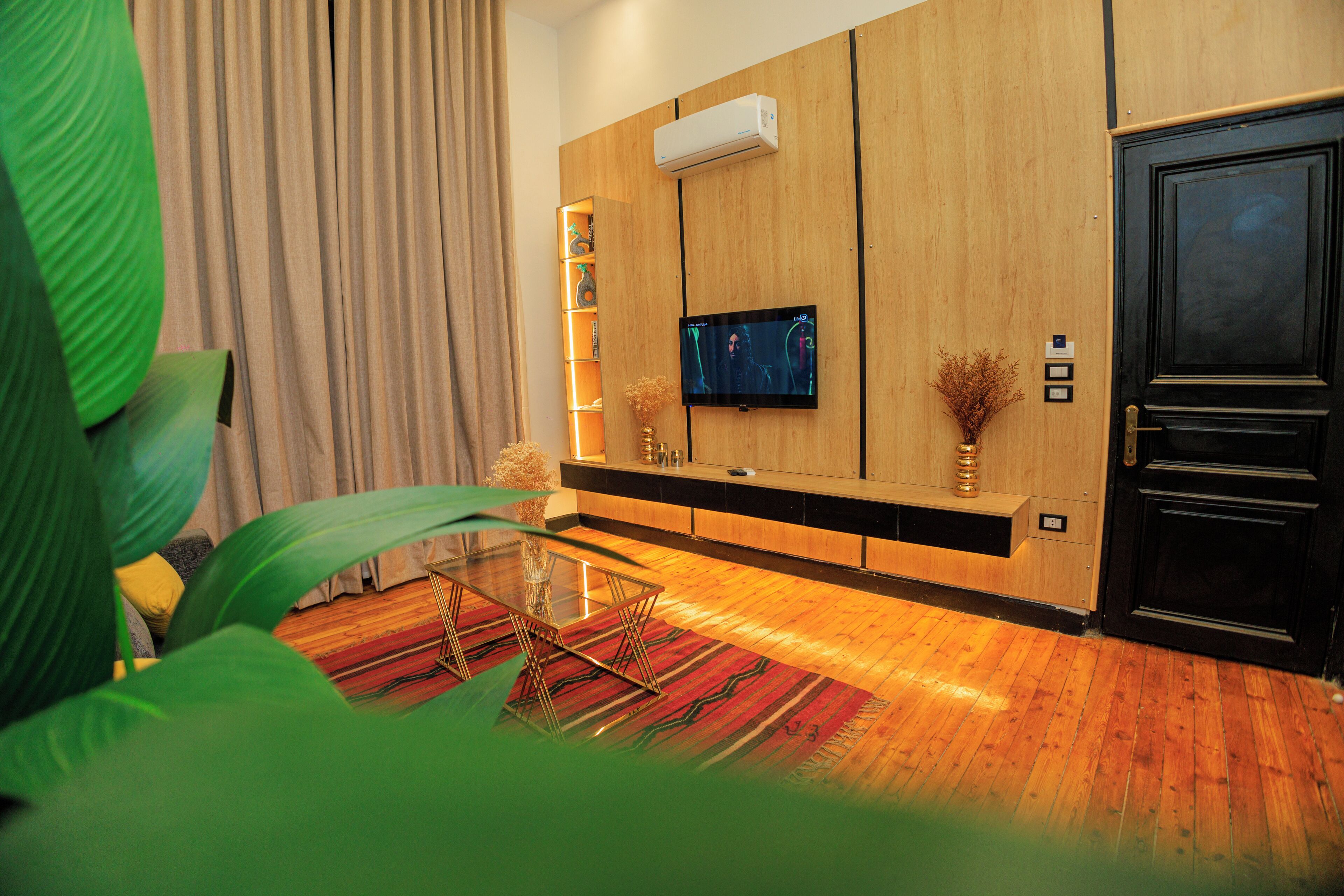 Una sala de estar moderna con un televisor de pantalla plana montado en una pared de madera, una mesa de centro de vidrio, un tapete a rayas rojo y negro, y una puerta negra.