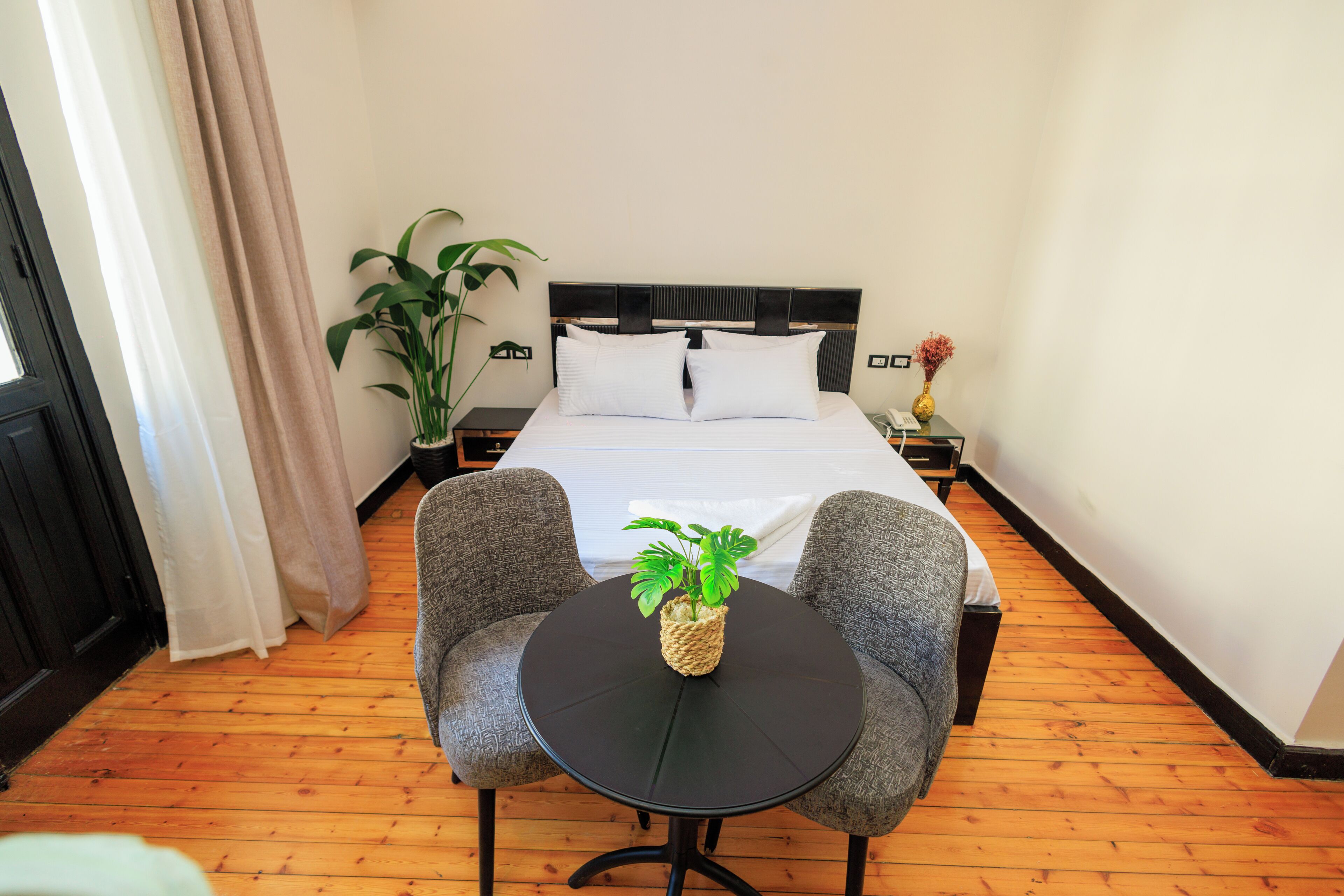 Een hotelkamer met een bed, twee fauteuils, een kleine ronde tafel met een plant, een bloempot en een nachtkastje met een vaas.