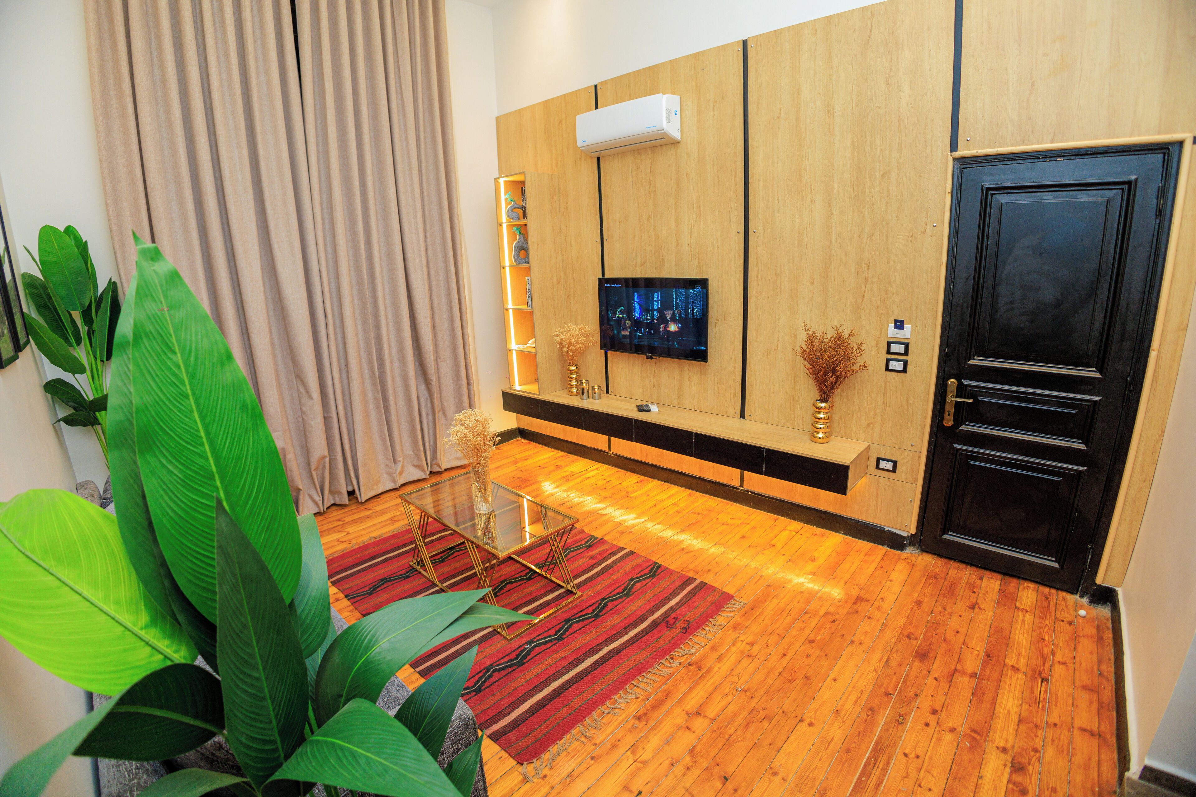 A modern living room with a wooden floor, a glass coffee table, a flat-screen TV mounted on the wall, and a black door.
