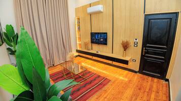 A modern living room with a wooden floor, a glass coffee table, a flat-screen TV mounted on the wall, and a black door.