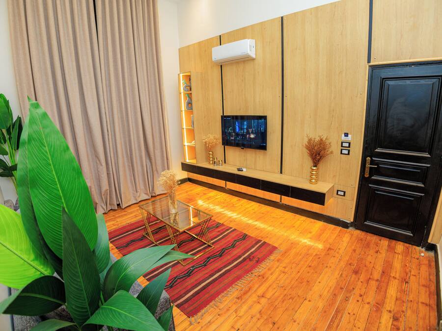 A modern living room with a wooden floor, a glass coffee table, a flat-screen TV mounted on the wall, and a black door.