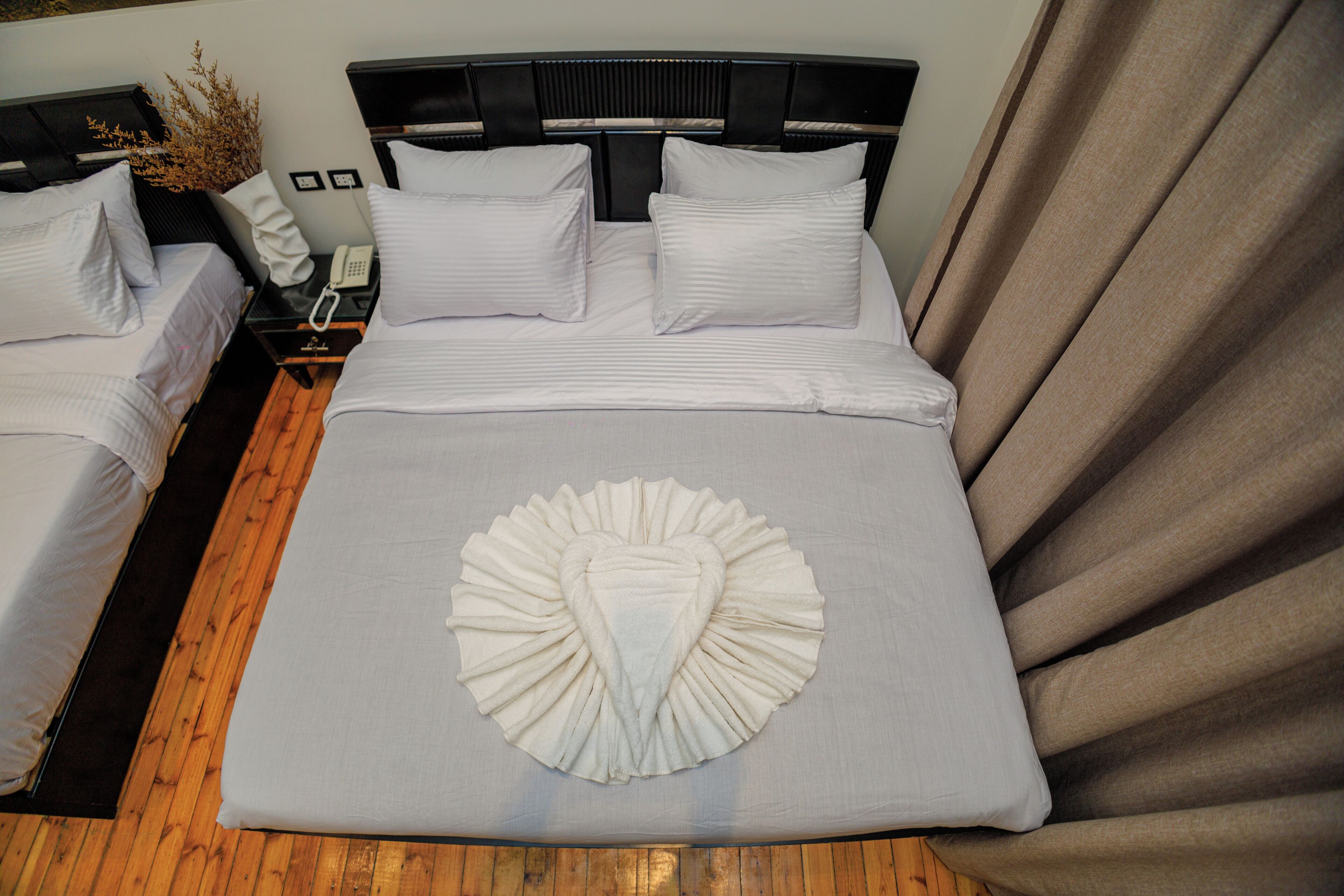 A hotel room with two beds, a wooden floor, a large decorative pillow, a telephone on the nightstand, and a curtain.